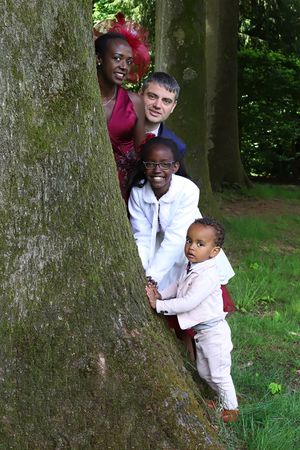 Family group poses playfully around large tree trunk in outdoor setting.