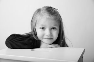 Child resting head on arm while posing at desk in black and white portrait.
