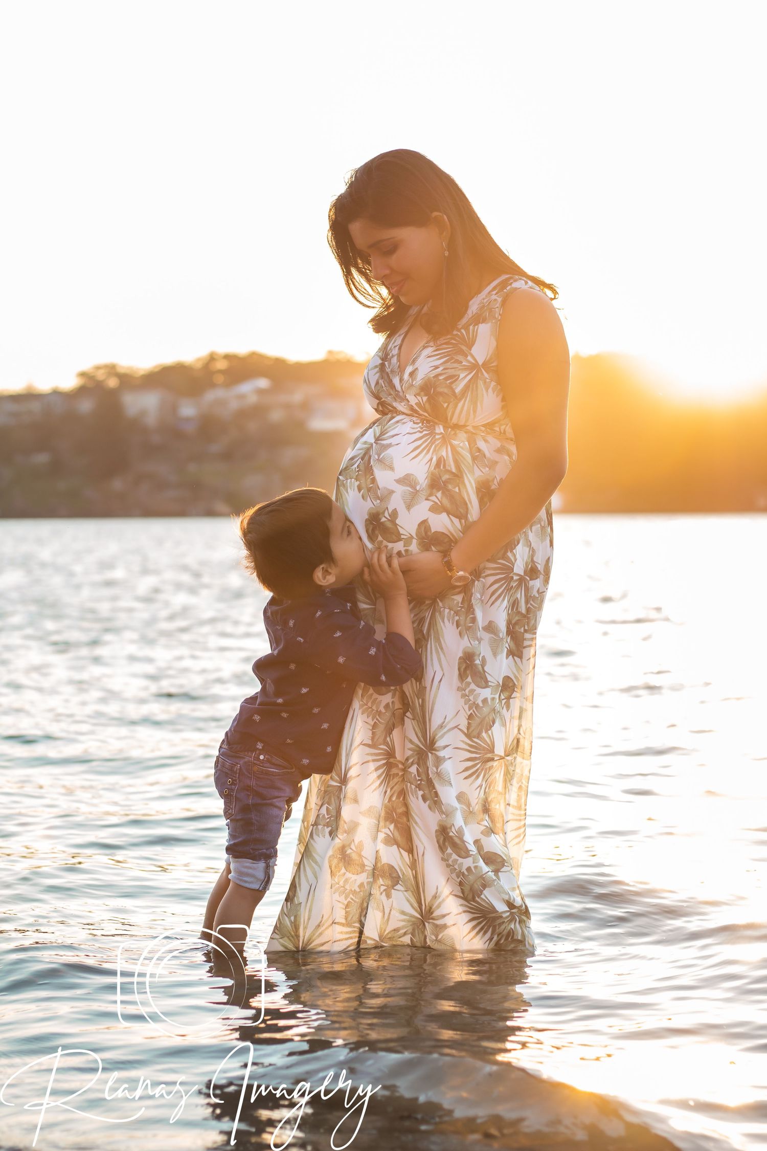 Pregnant mother and toddler share a tender moment at sunset by the water.
