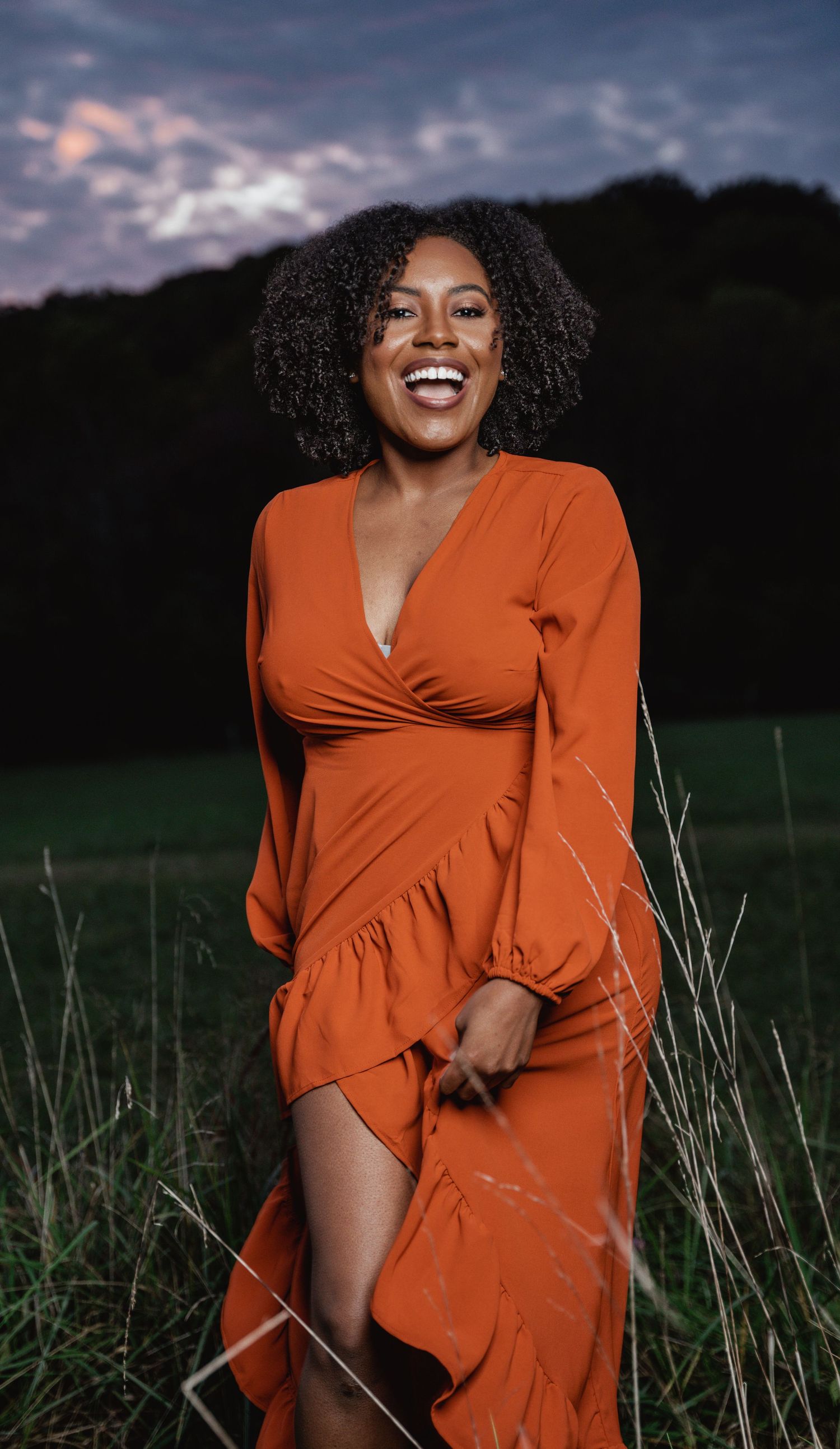 Joyful woman in orange ruffle dress standing in grassy field at sunset.