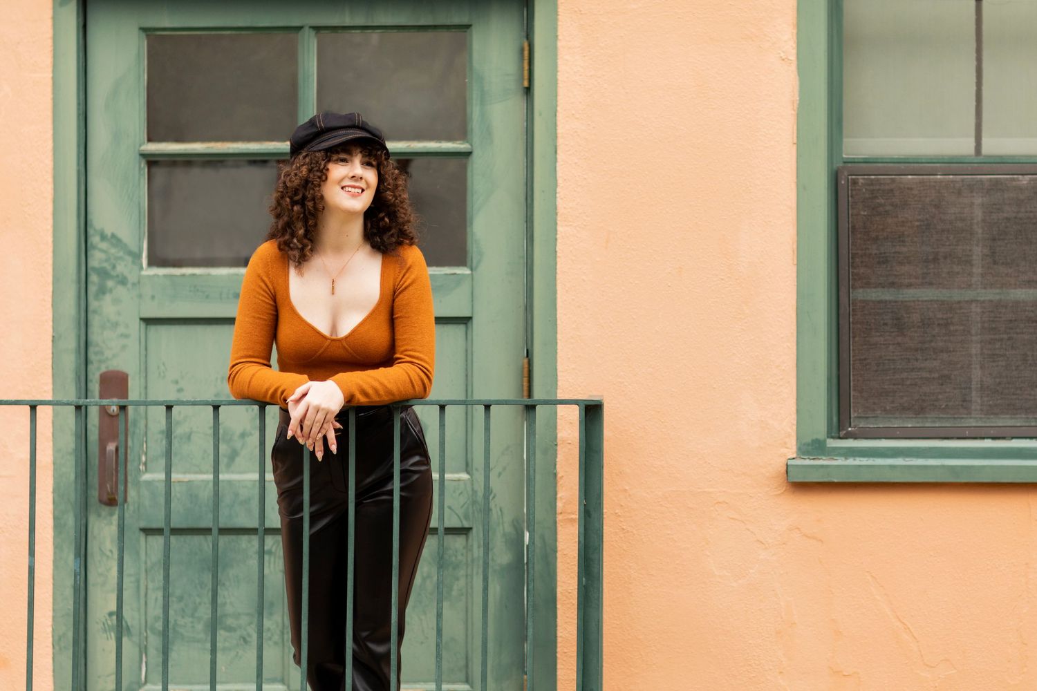 A high school senior in orange sweater and black pants poses on green balcony against peach colored wall.