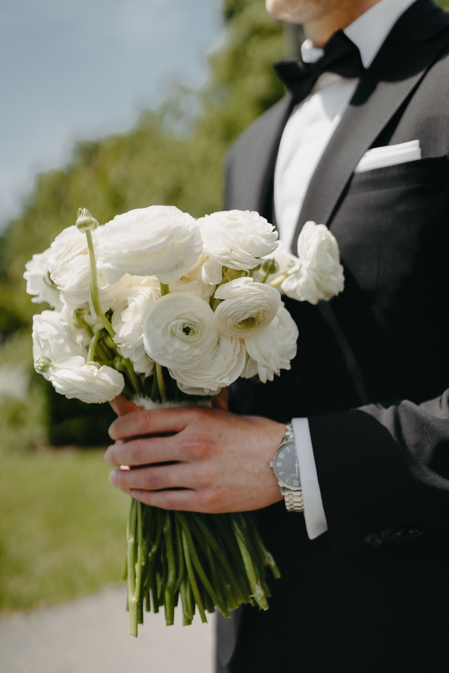 A bouquet of white ranunculus flowers held by hands in a black tuxedo sleeve.