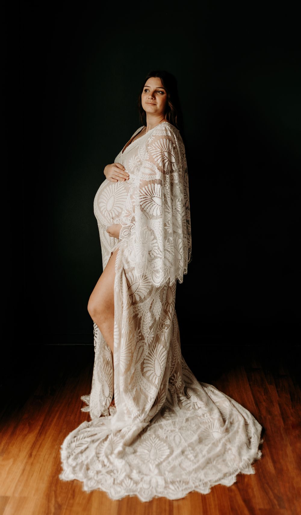 A dramatic maternity portrait in a flowing lace gown against dark backdrop.