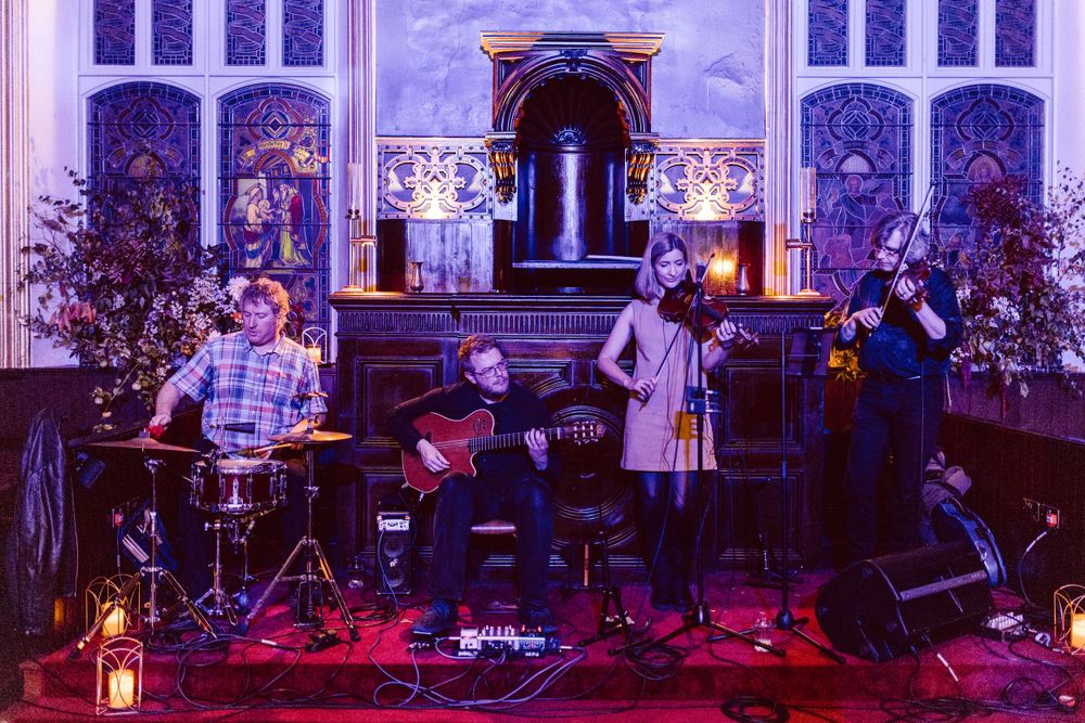 Band performs on stage in a church setting with purple lighting and gothic architecture.