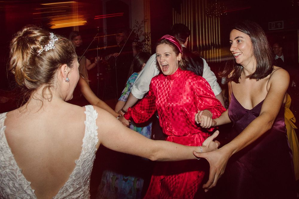 Wedding guests dancing energetically in red and dark dresses on a dimly lit dance floor.