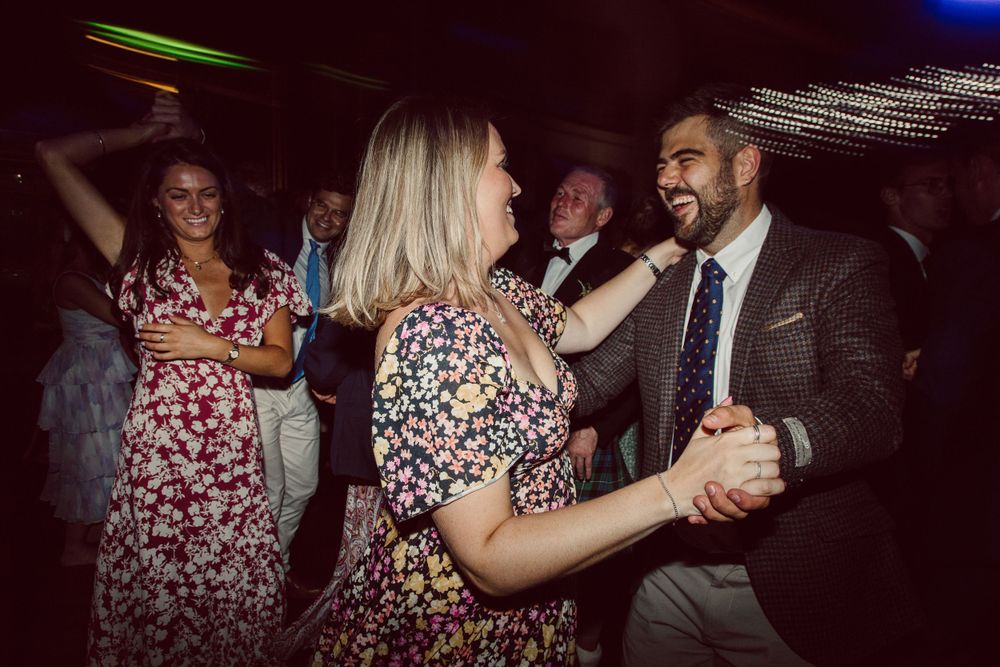 Wedding guests dance together at a lively reception wearing floral dresses and formal attire.