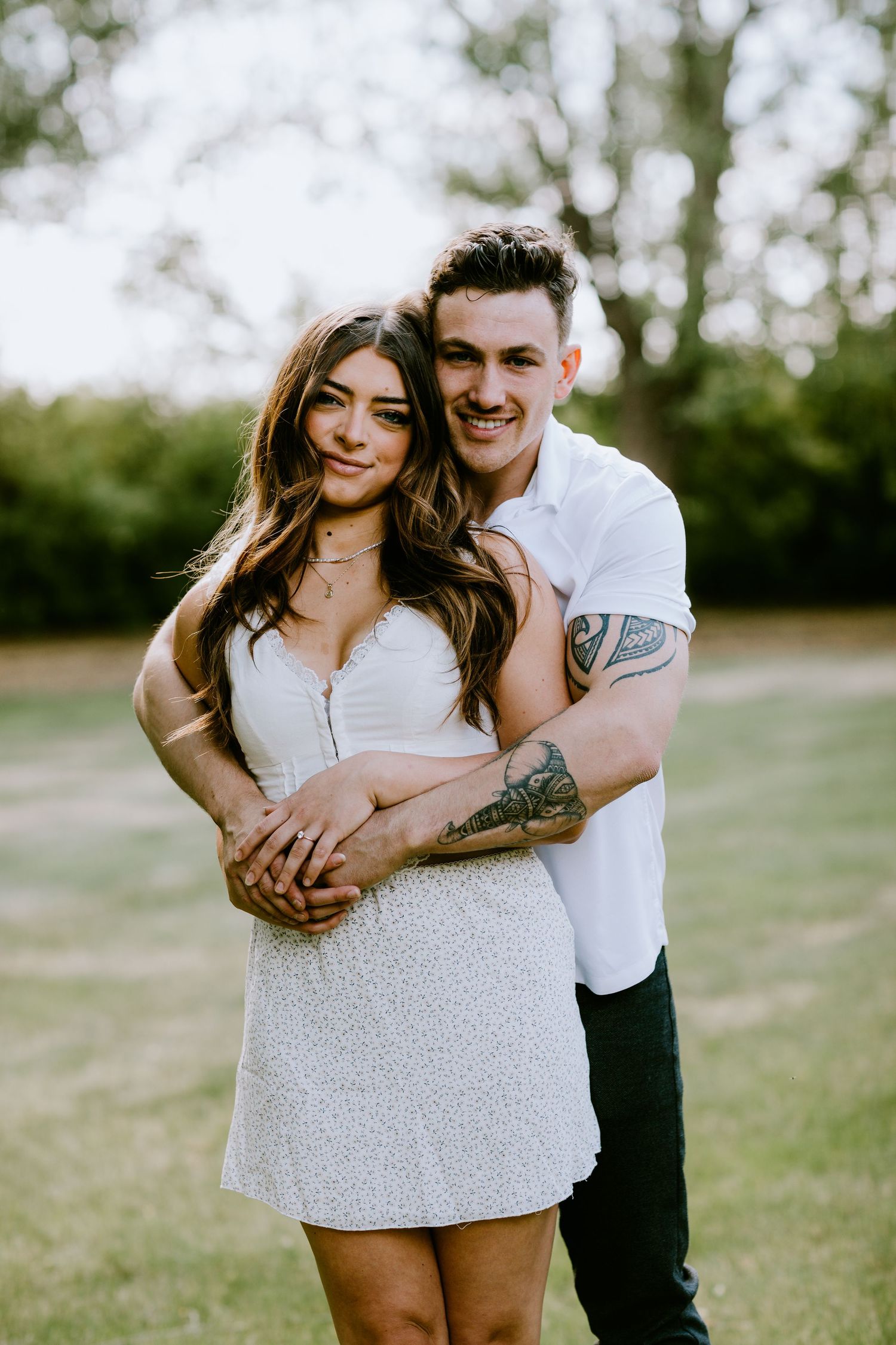 A couple poses together outdoors with the person in white embracing from behind.