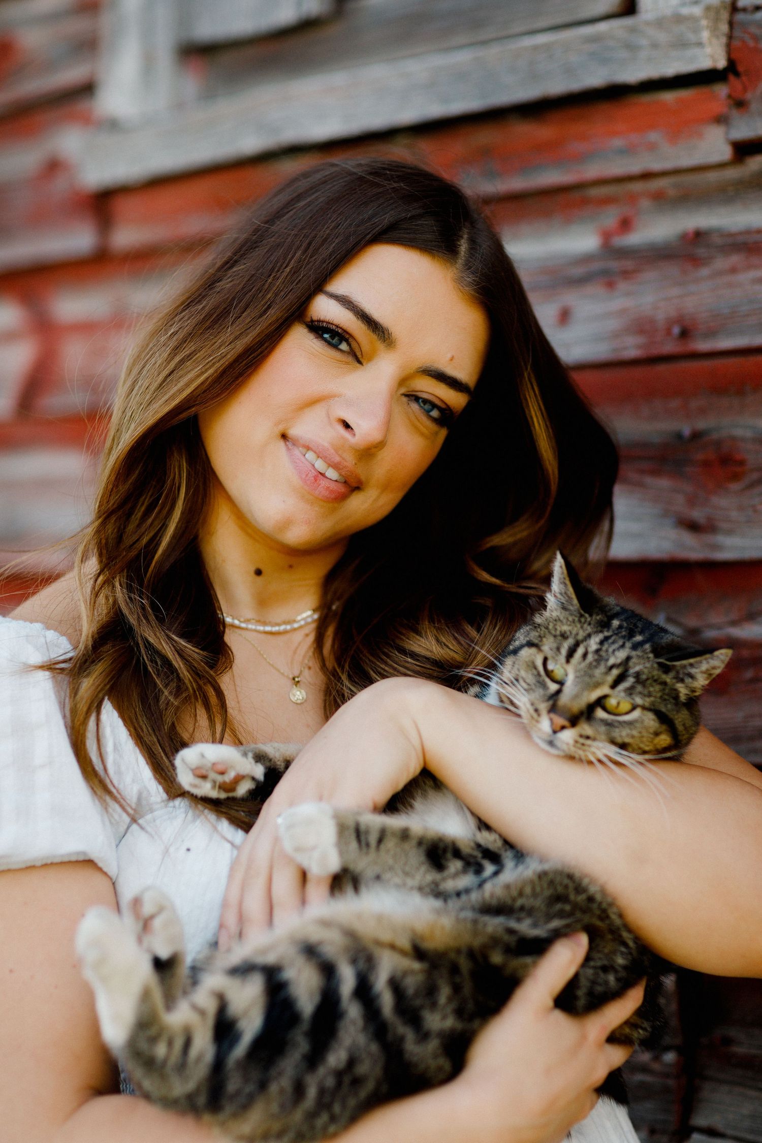 A woman in white holds a contented tabby cat while sitting by a rustic wall.