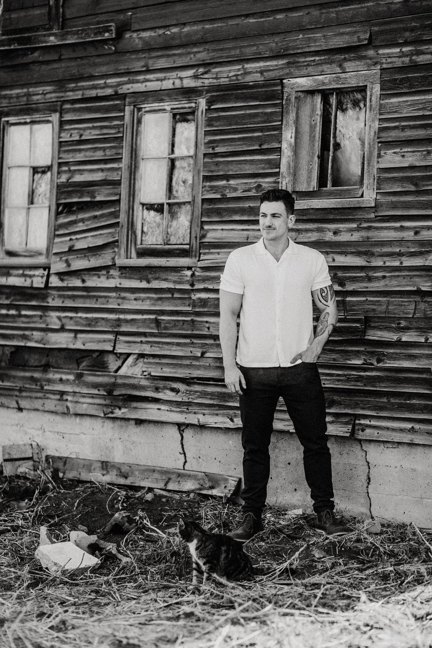 A person in white stands against a weathered wooden building with a cat nearby.