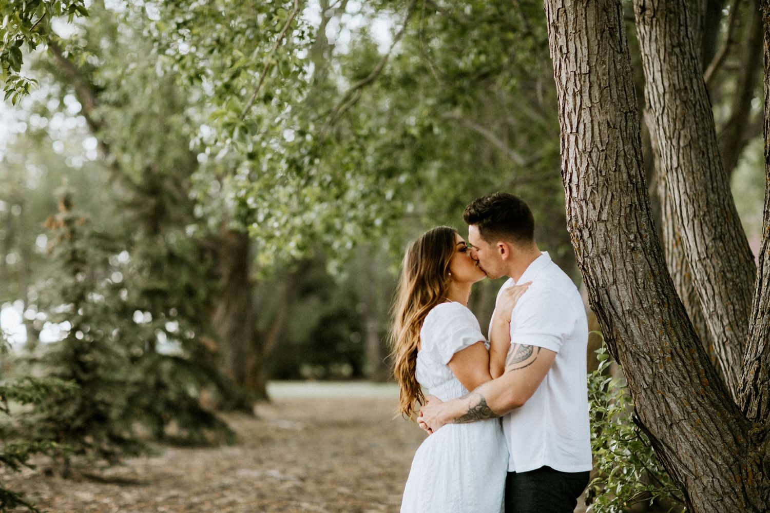Romantic couple in white embracing on tree-lined path during outdoor photoshoot.