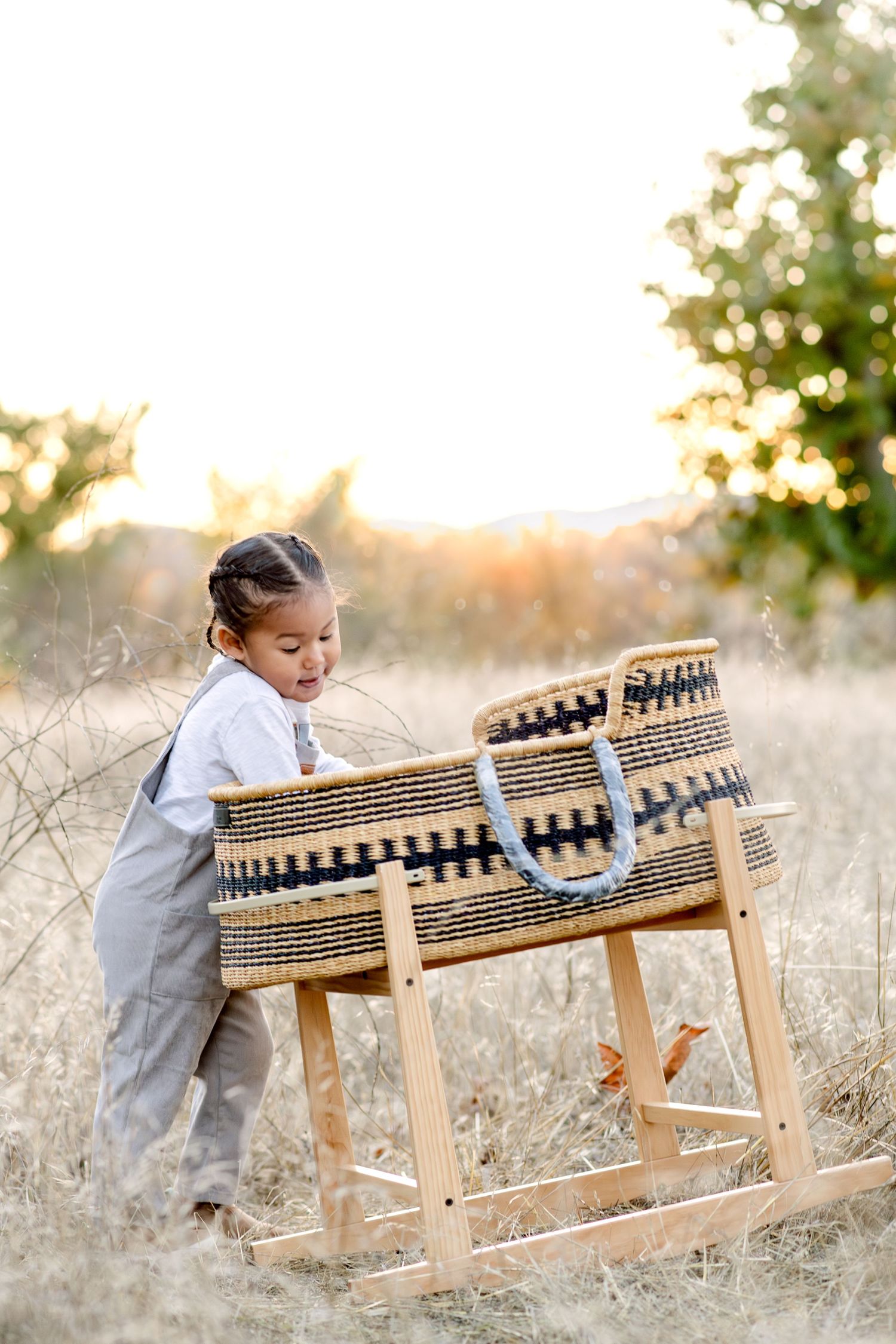Child playing with wooden rocking cradle in sunlit field with natural decor.