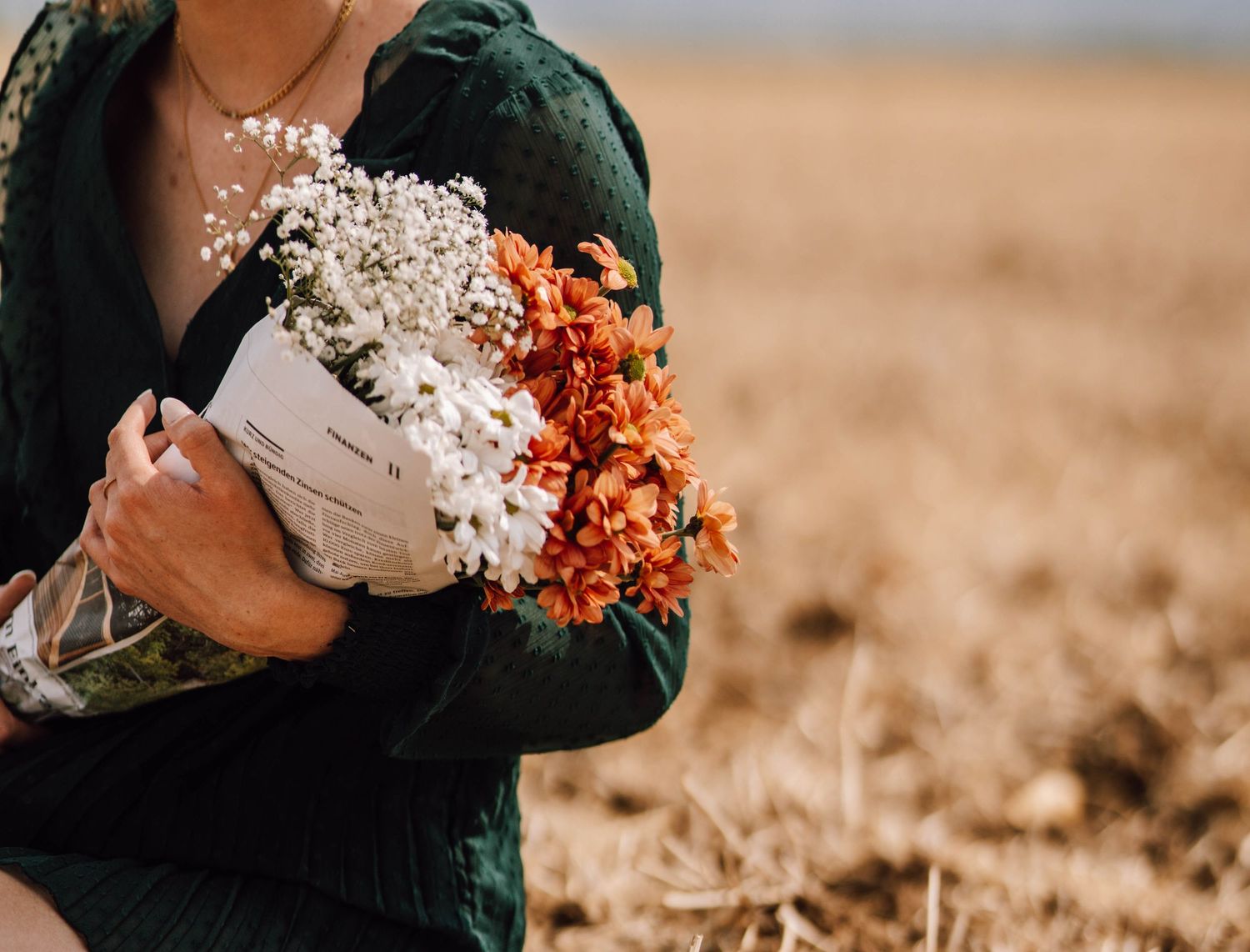 Hands holding a bouquet of white and orange flowers against a rustic field backdrop.