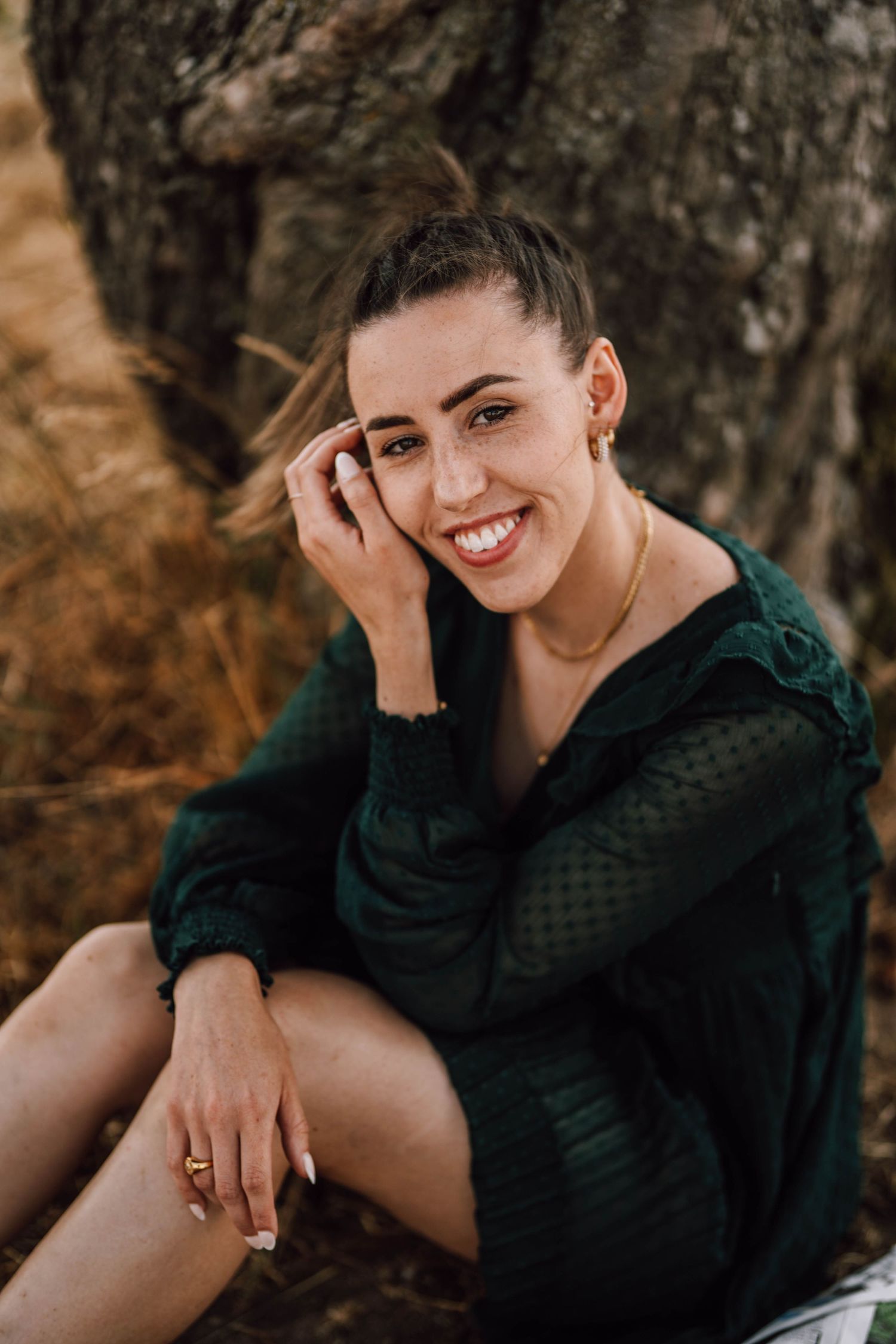 A person in a dark green dress poses casually against a tree trunk.