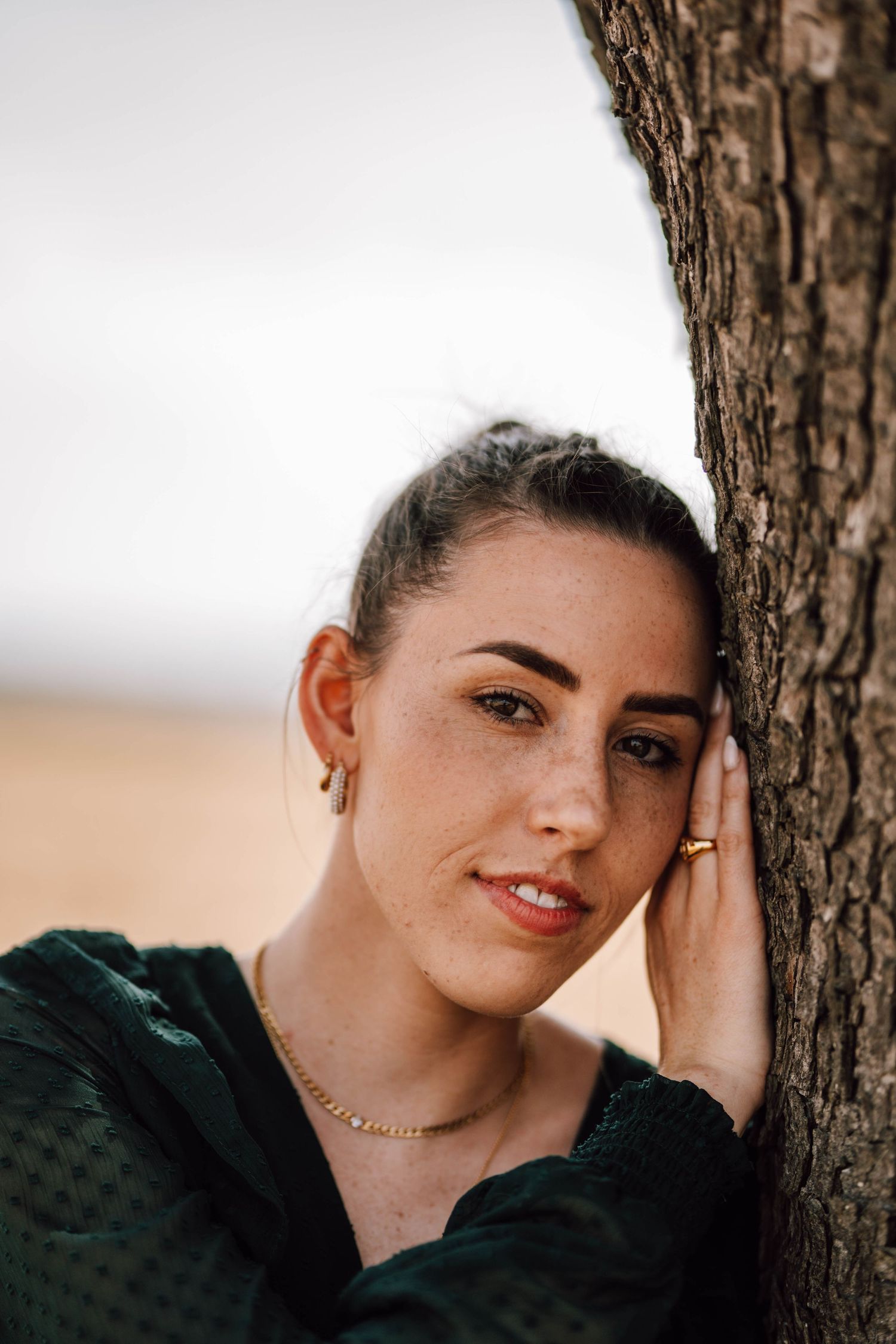 A portrait of someone leaning against a tree trunk wearing a dark green top.