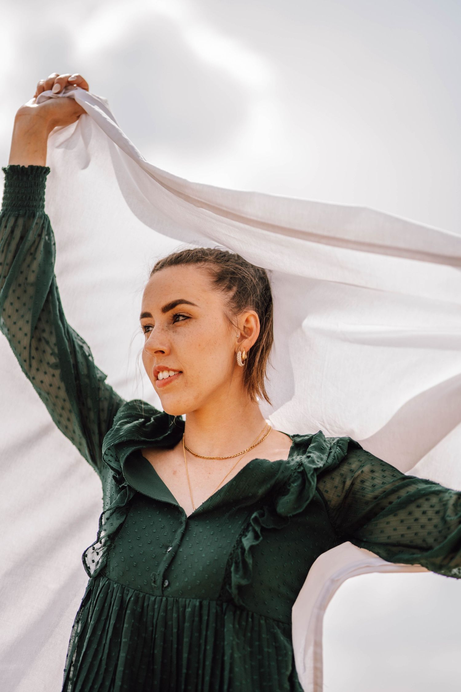 A person in a green dress holds up a flowing white fabric against a bright sky.