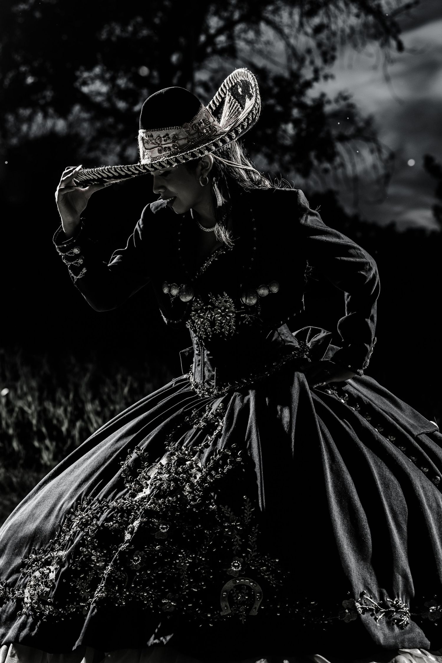 A dramatic black and white photograph of a person in an ornate Victorian-style dress and sombrero dancing.