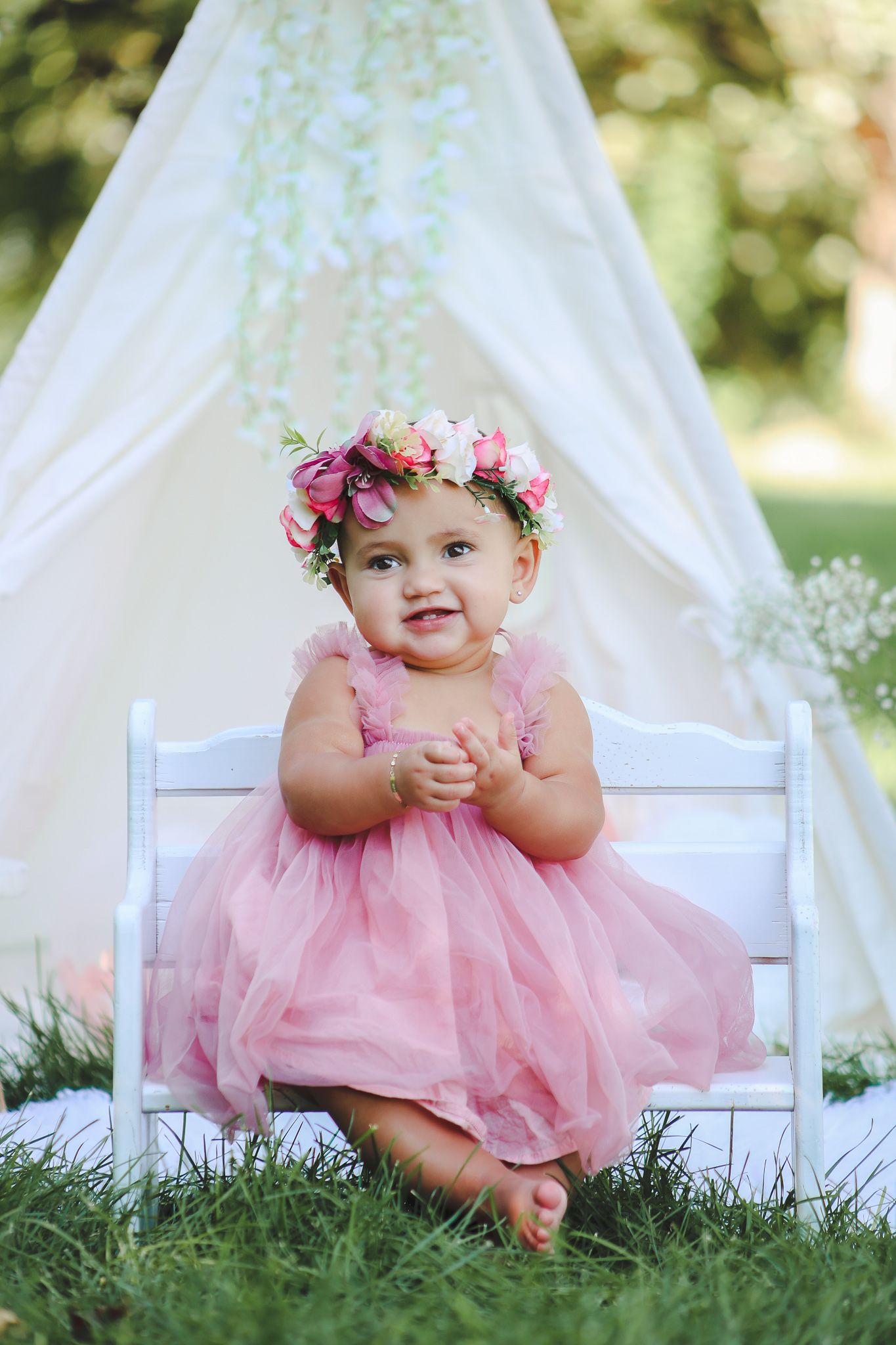 A baby in a pink tulle dress sits on a white chair outdoors with a floral crown and dreamy white tent backdrop.