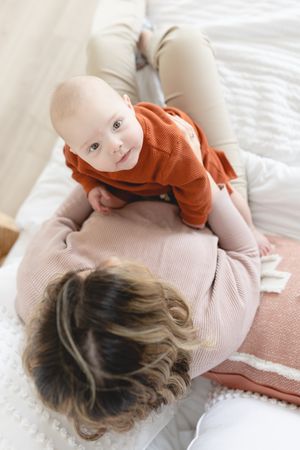 Baby in orange sweater lies on a parent's lap on white bedding in a soft, natural light setting.