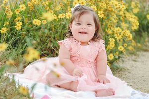 Smiling toddler in pink dress sitting on blanket surrounded by yellow wildflowers.