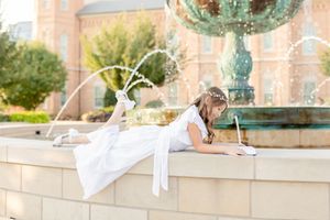 Young dancer in a white dress performs a graceful pose on a stone wall near an ornate fountain in an urban setting.