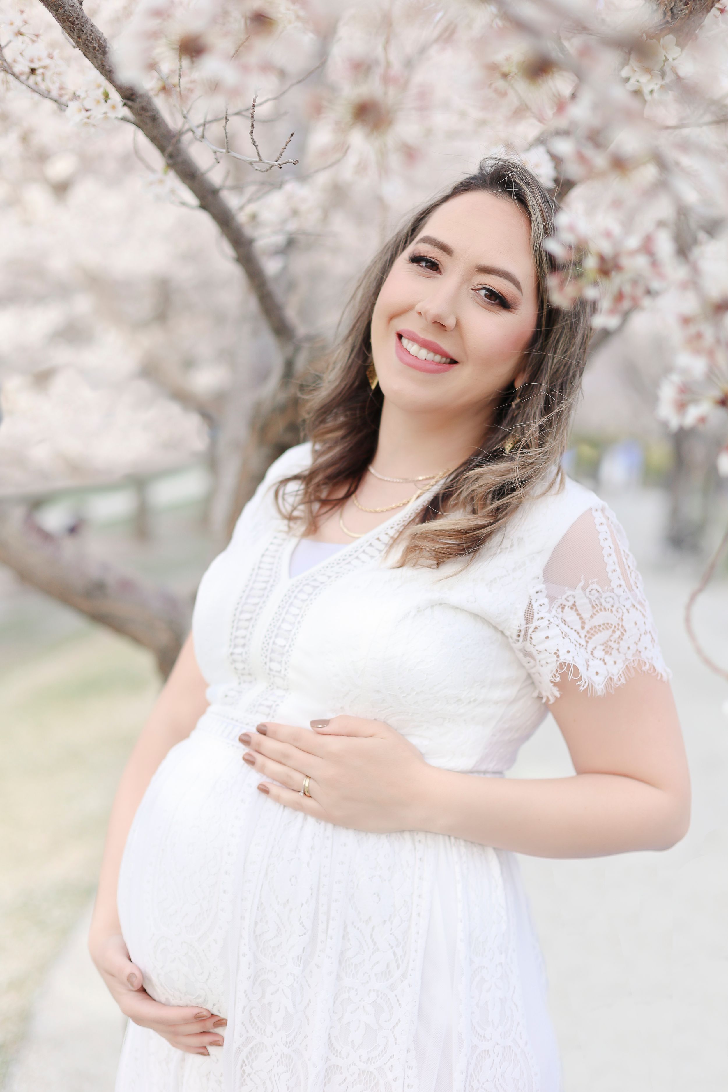 Elegant maternity portrait under blooming cherry blossoms with a white lace dress in a dreamy spring setting.