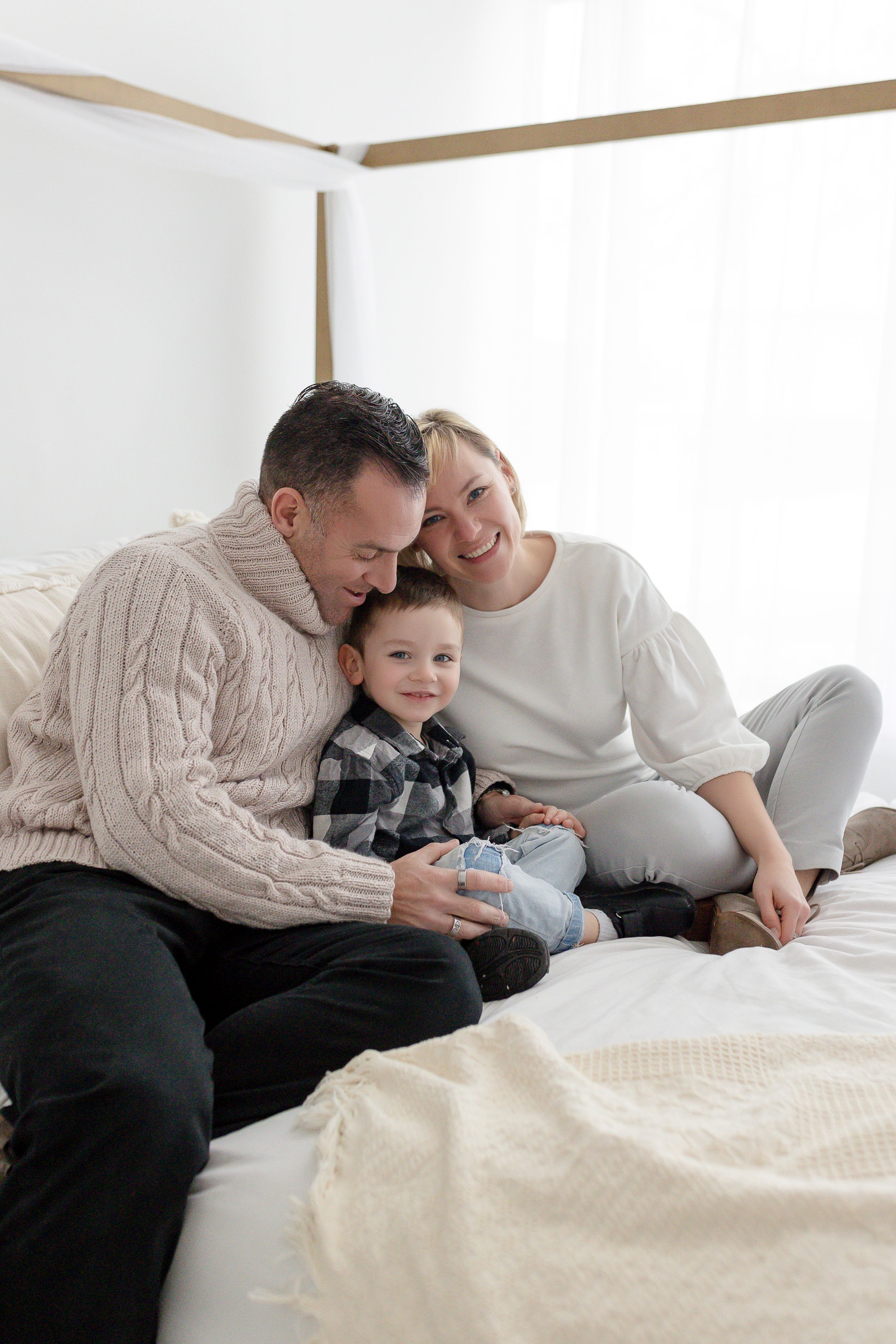 A happy family shares a tender moment together while relaxing on a white bed in casual sweaters and clothing.