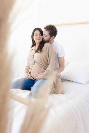 A tender intimate moment between expectant parents sitting together on a white bed in soft natural lighting.