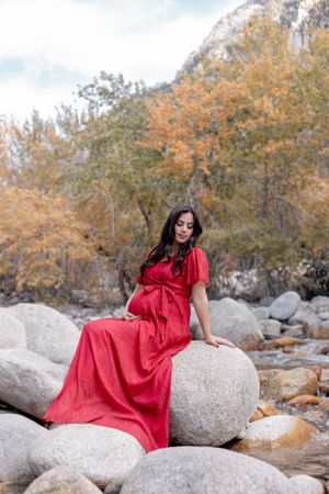 A person in a flowing red dress sits gracefully on large river rocks surrounded by autumn trees and mountain scenery.