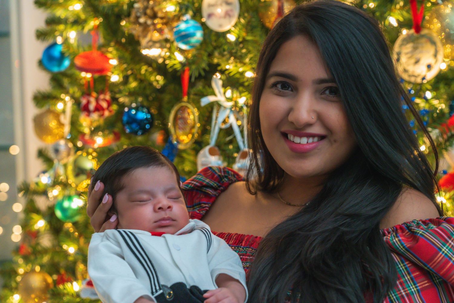A newborn baby in striped outfit being held in front of a decorated Christmas tree with ornaments.