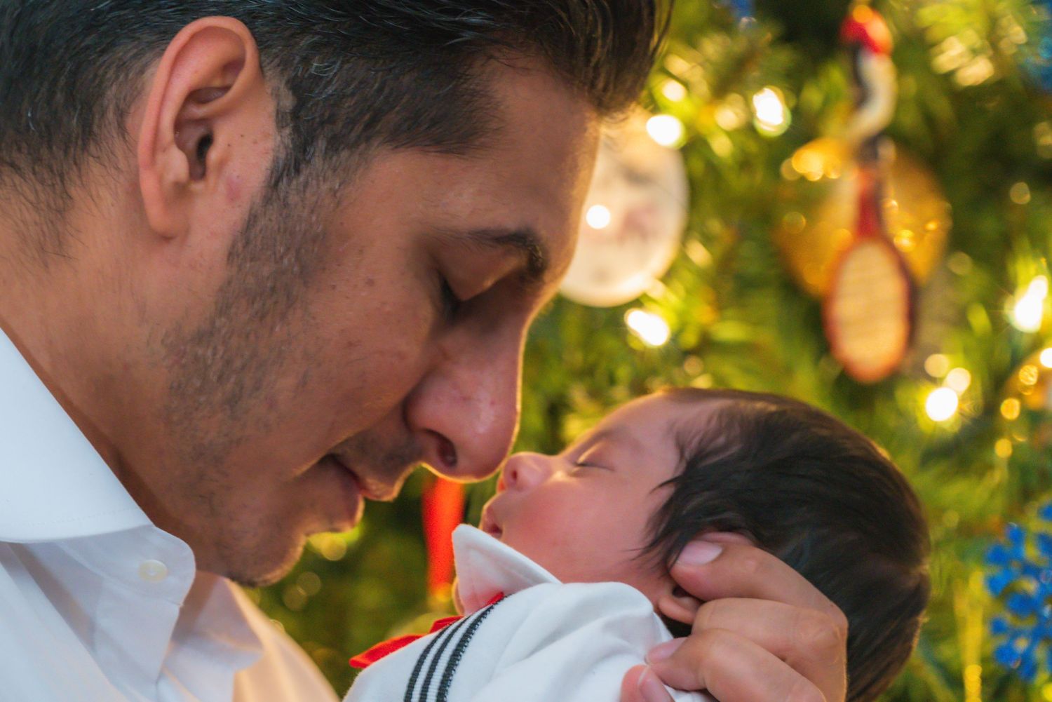 A tender moment between parent and newborn baby in front of a glowing Christmas tree with bokeh lights.