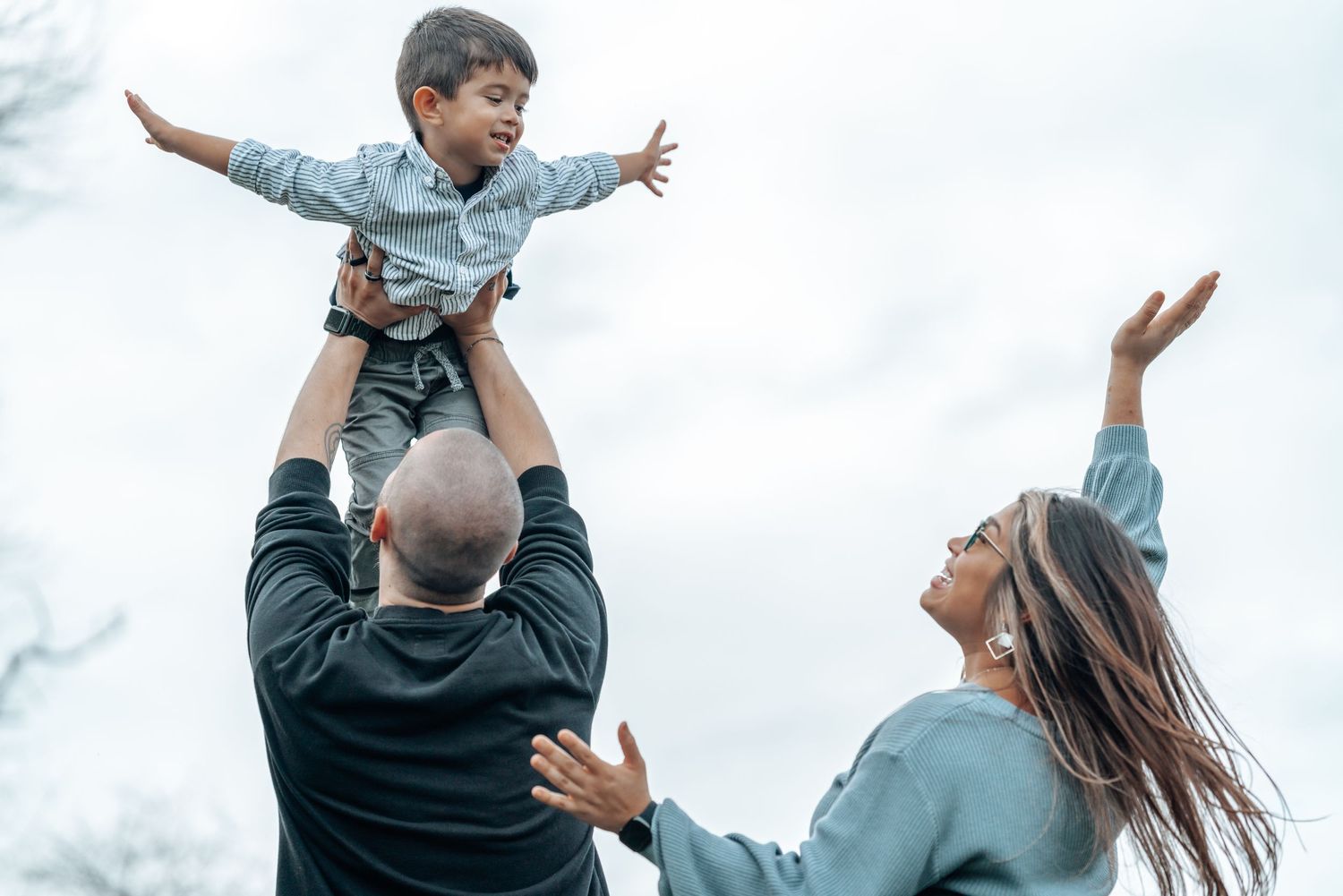 Playful family moment with child being lifted high in the air against cloudy sky.