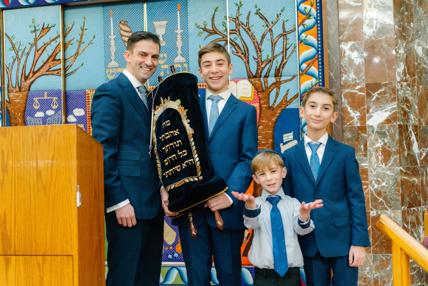 A group in matching navy suits poses with a Torah scroll against a colorful synagogue backdrop.