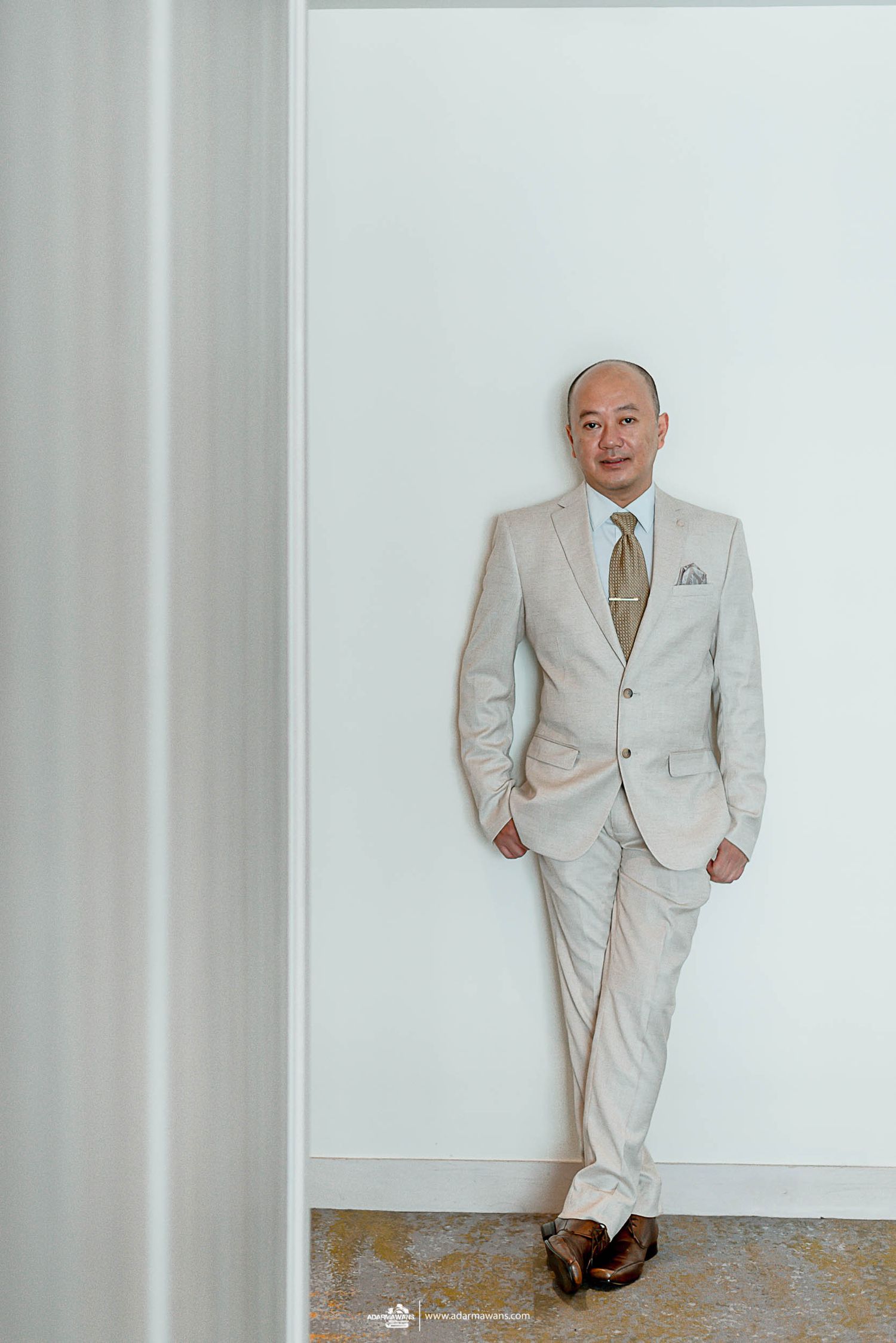A groom in a light beige suit leans casually against a white wall.