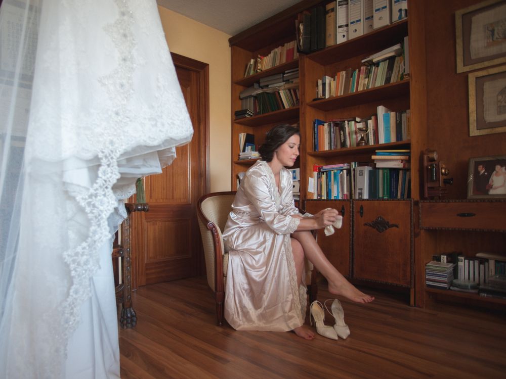 Bride in silk robe sitting quietly beside her wedding dress in a book-lined room.