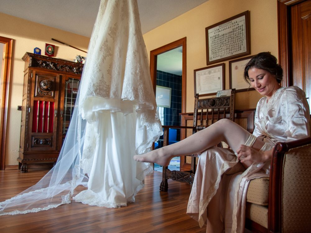 Bride in robe putting on stockings beside her hanging lace wedding gown indoors.