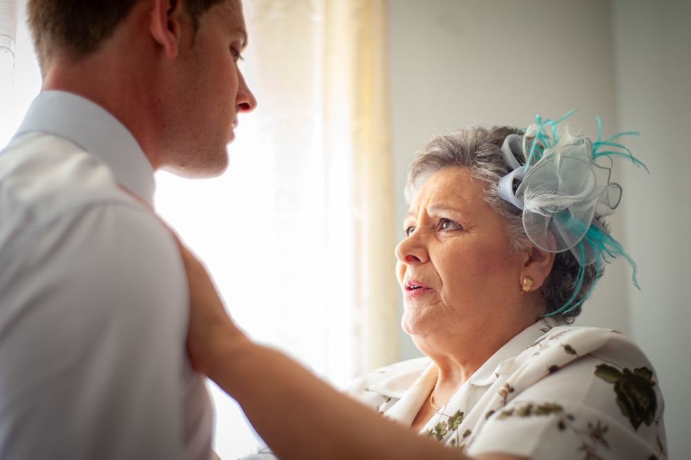 Older woman in floral dress and teal fascinator lovingly adjusts the groom's shirt before the wedding.