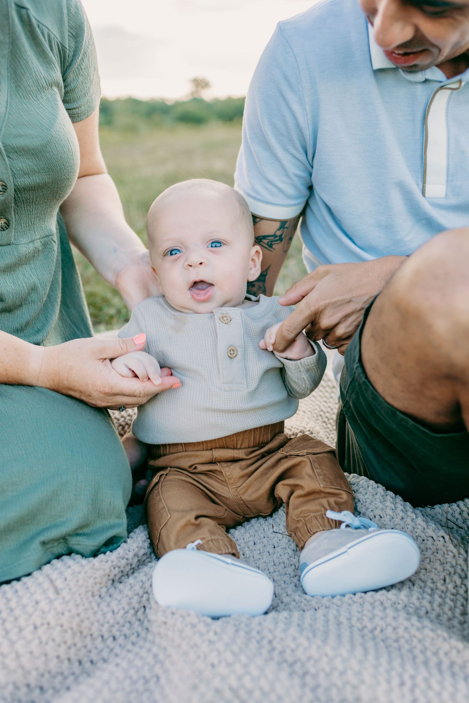 Baby sitting on blanket outdoors supported by parents during casual photo session.