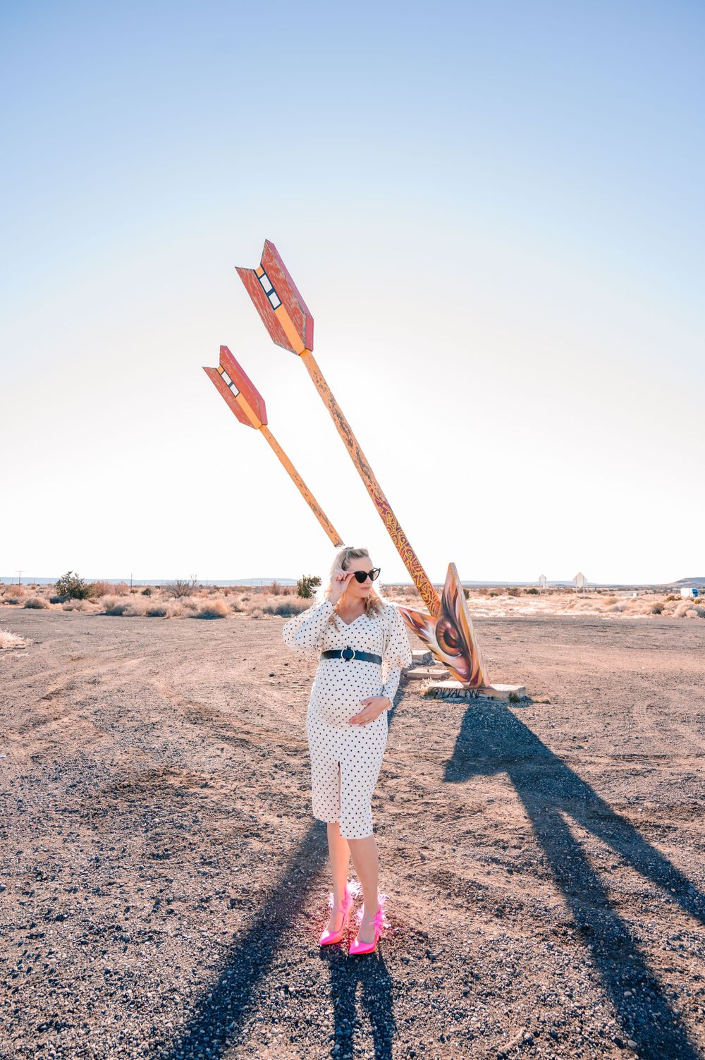 Woman in vintage-style dress stands against desert backdrop with large orange arrows.