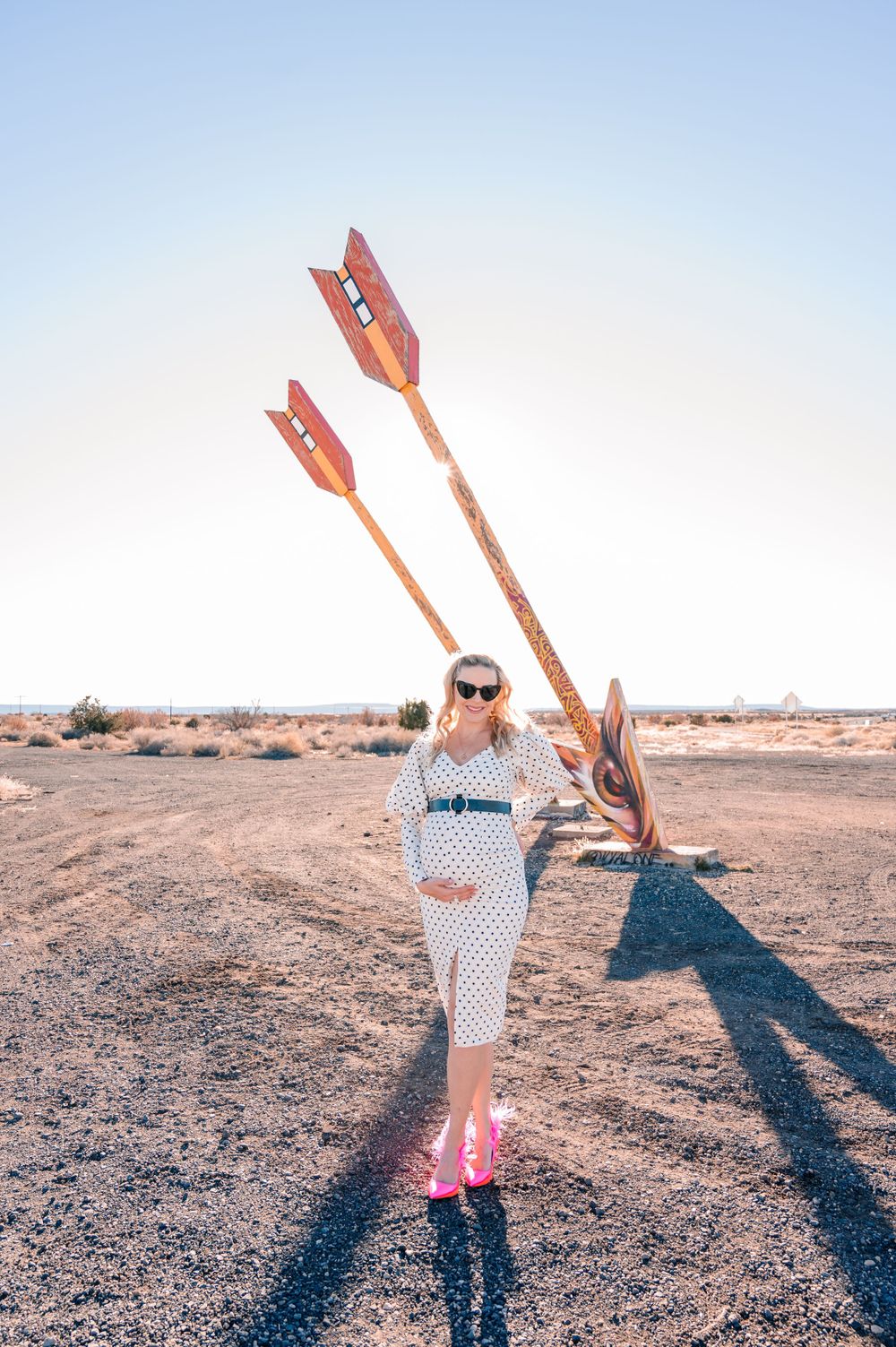 Woman in polka dot dress poses with giant decorative arrows in desert landscape.