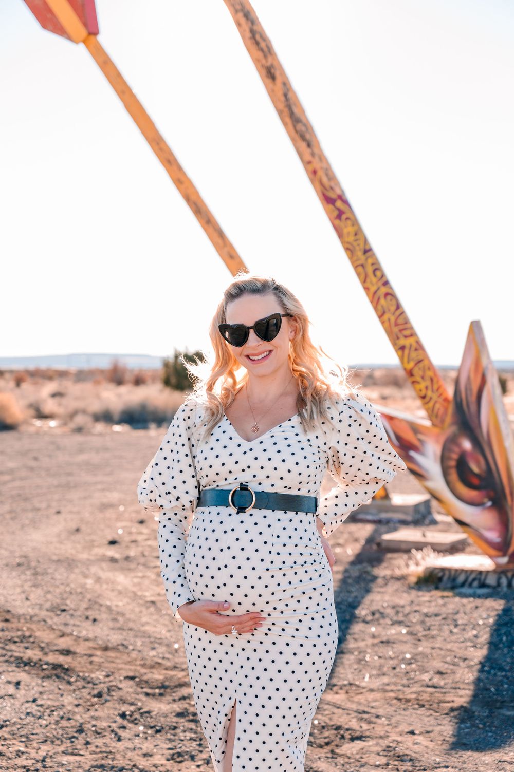 Woman in polka dot maternity dress poses with decorative arrows in desert setting.