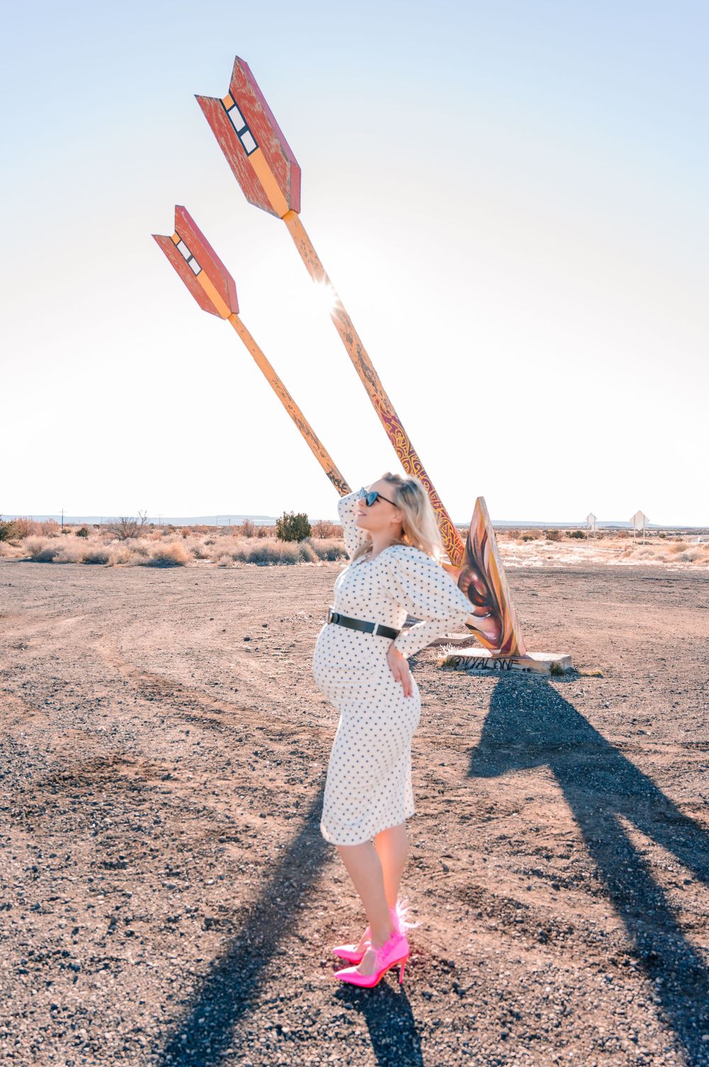 Woman in sunglasses poses playfully with large orange arrows in desert environment.