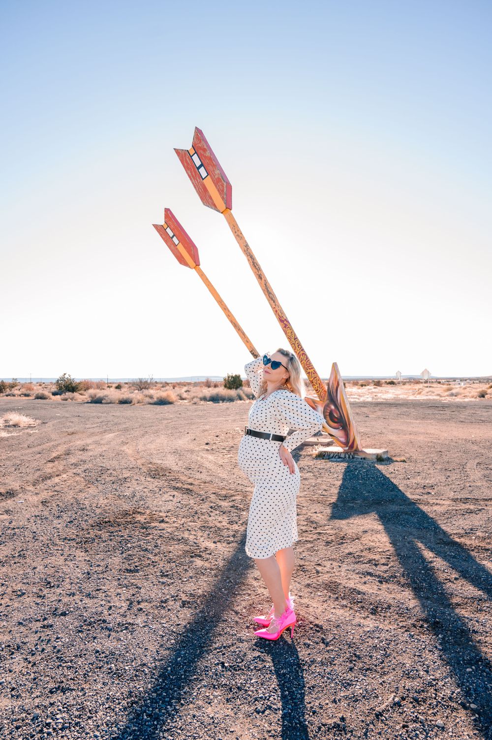 Woman in retro dress and pink shoes poses with giant arrows in desert setting.