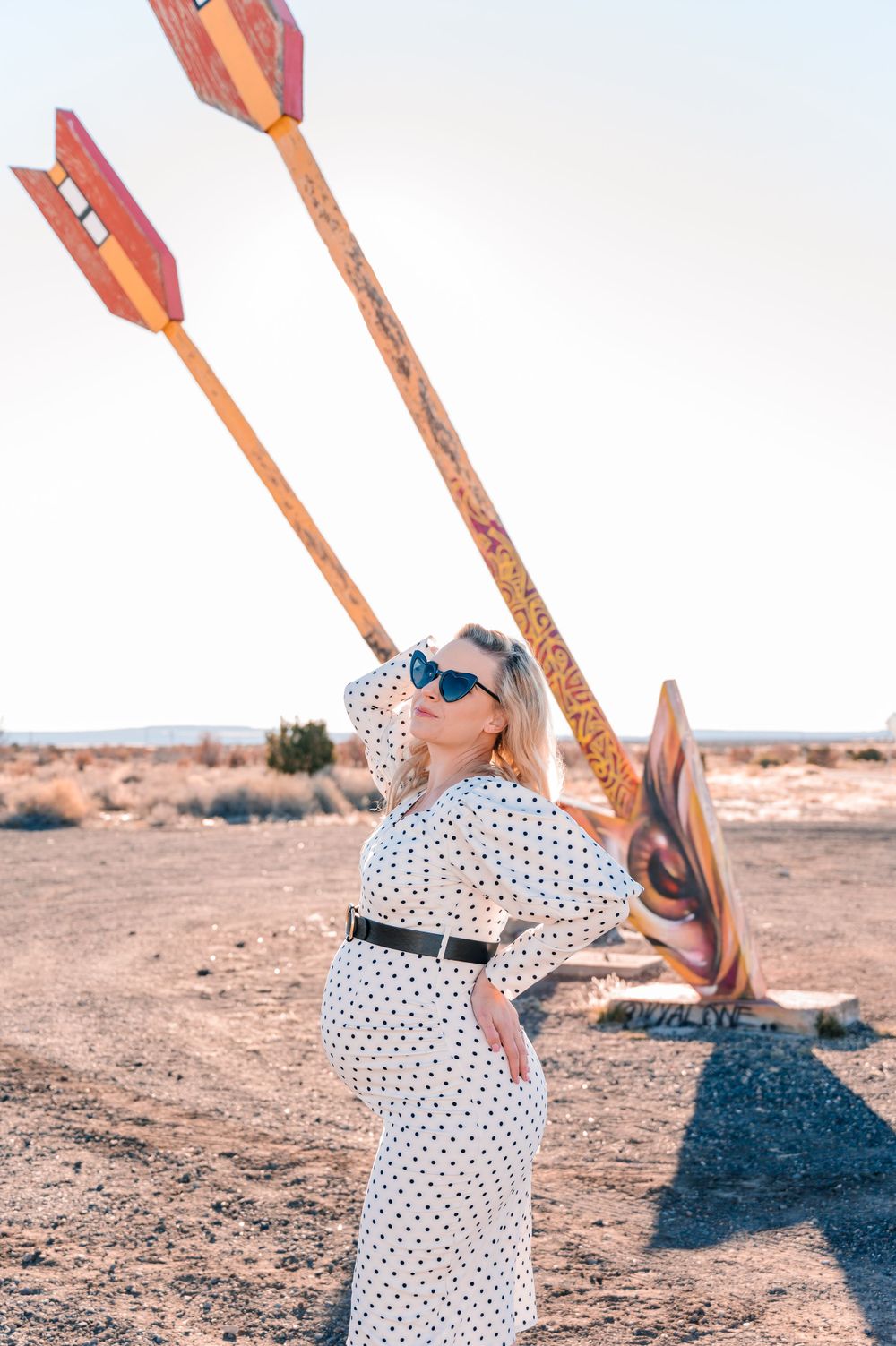 Woman in polka dot dress poses beside large orange arrow sculptures in desert landscape.