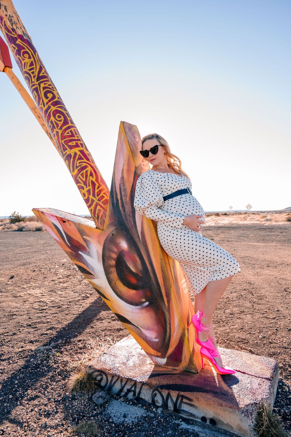 Woman poses on decorative arrow sculpture in desert with bright pink shoes.