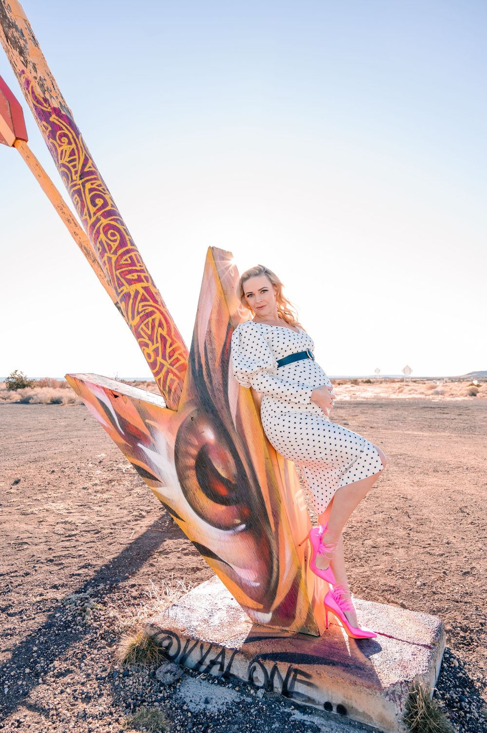 Woman in vintage dress poses on Royal Love arrow sculpture in desert setting.