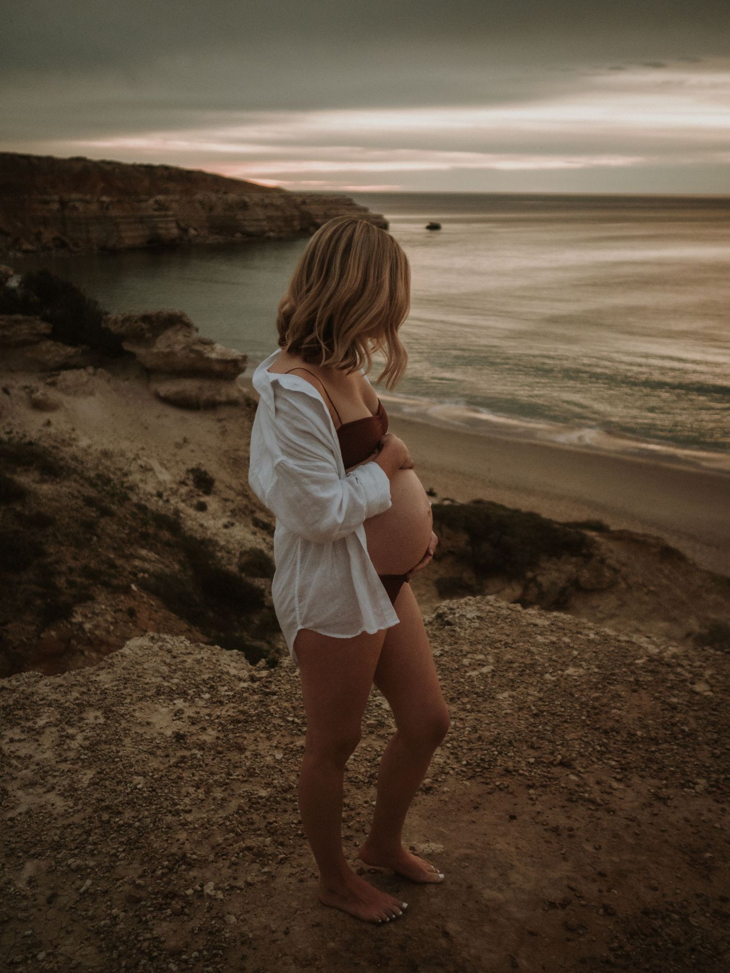 Woman in white shirt standing on coastal cliff during moody sunset at beach.