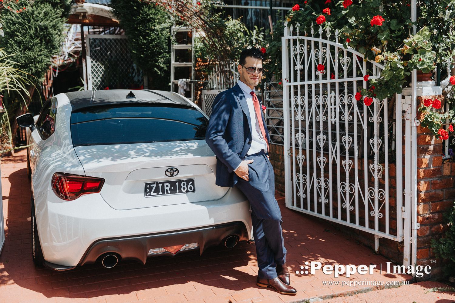 Groom in blue suit leaning against a white Toyota 86 sports car beside a rose-covered gate.