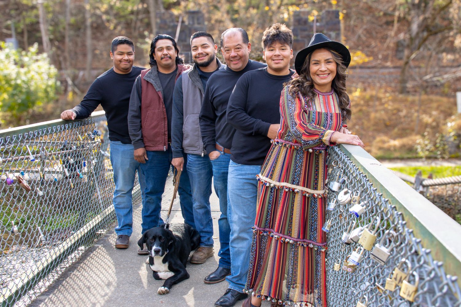 A group of six people and a dog pose together on a bridge with padlocks, surrounded by nature.