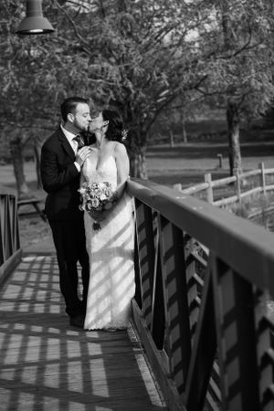 Black and white photo of bride and groom kissing on a wooden bridge surrounded by trees.