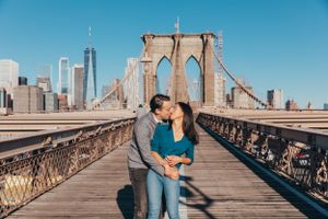 A couple sharing a kiss on the Brooklyn Bridge with the New York City skyline and blue sky behind them.